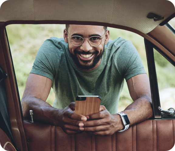 Homem sorrindo segurando um celular, apoiado na janela do carro